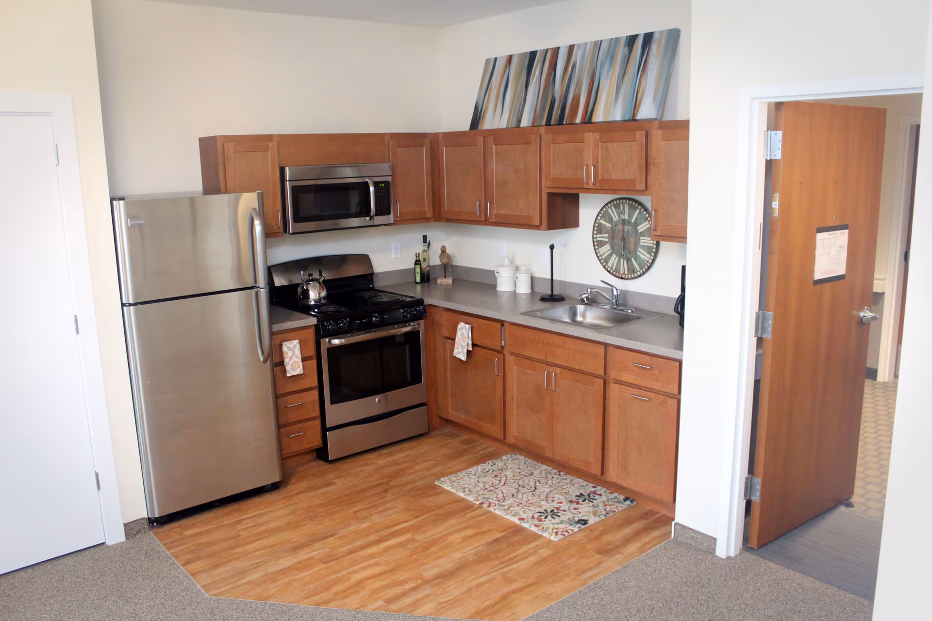 A compact kitchen area with wooden cabinets, a stainless steel refrigerator, stove, and microwave. The countertop has a sink, a few decorative items, and a large wall clock. There is a small patterned rug on the wooden floor section, and an open door leads to another room with carpeted flooring.