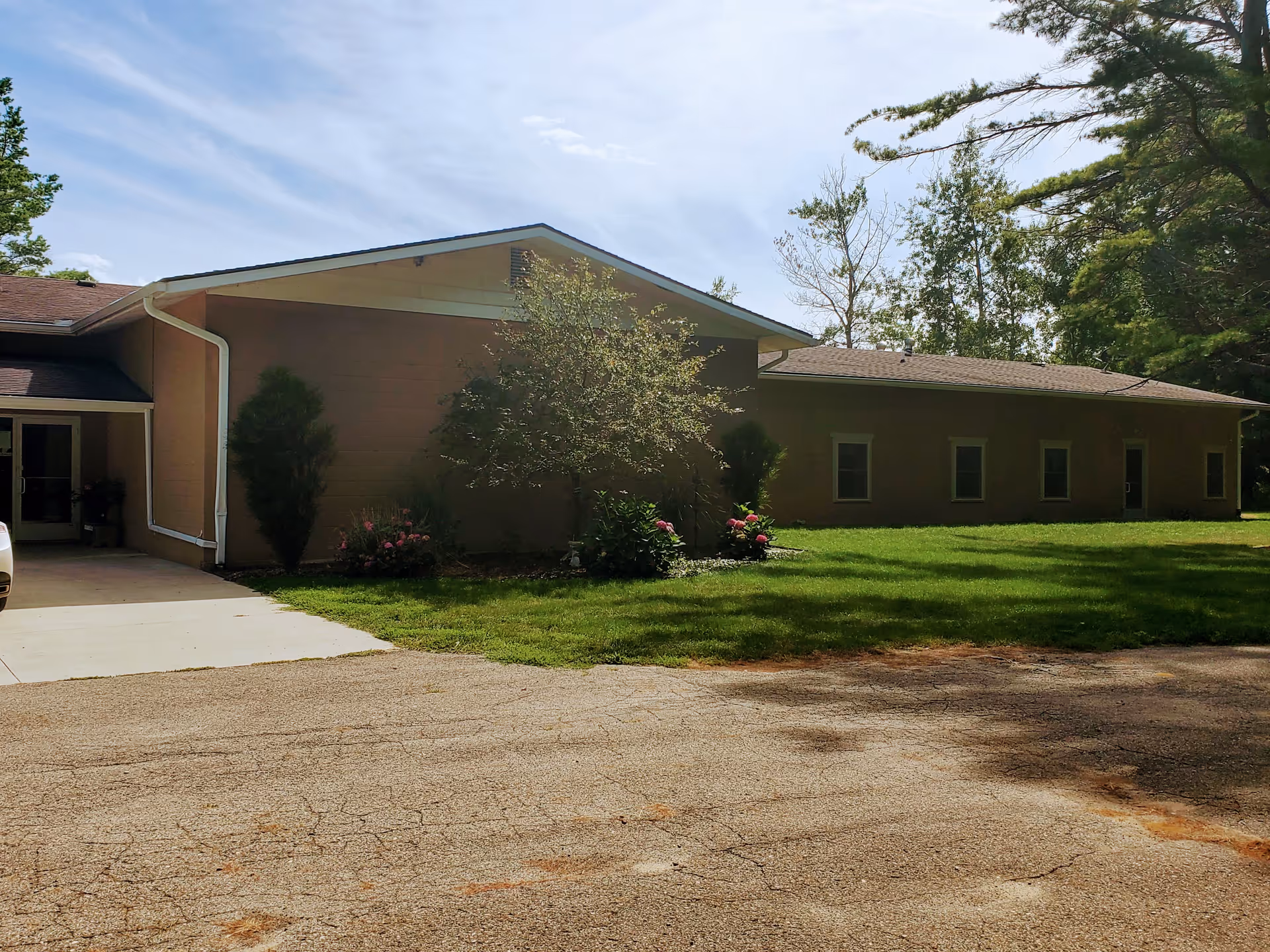 Single-story building with a driveway, lawn, shrubs and trees under a partly cloudy sky.