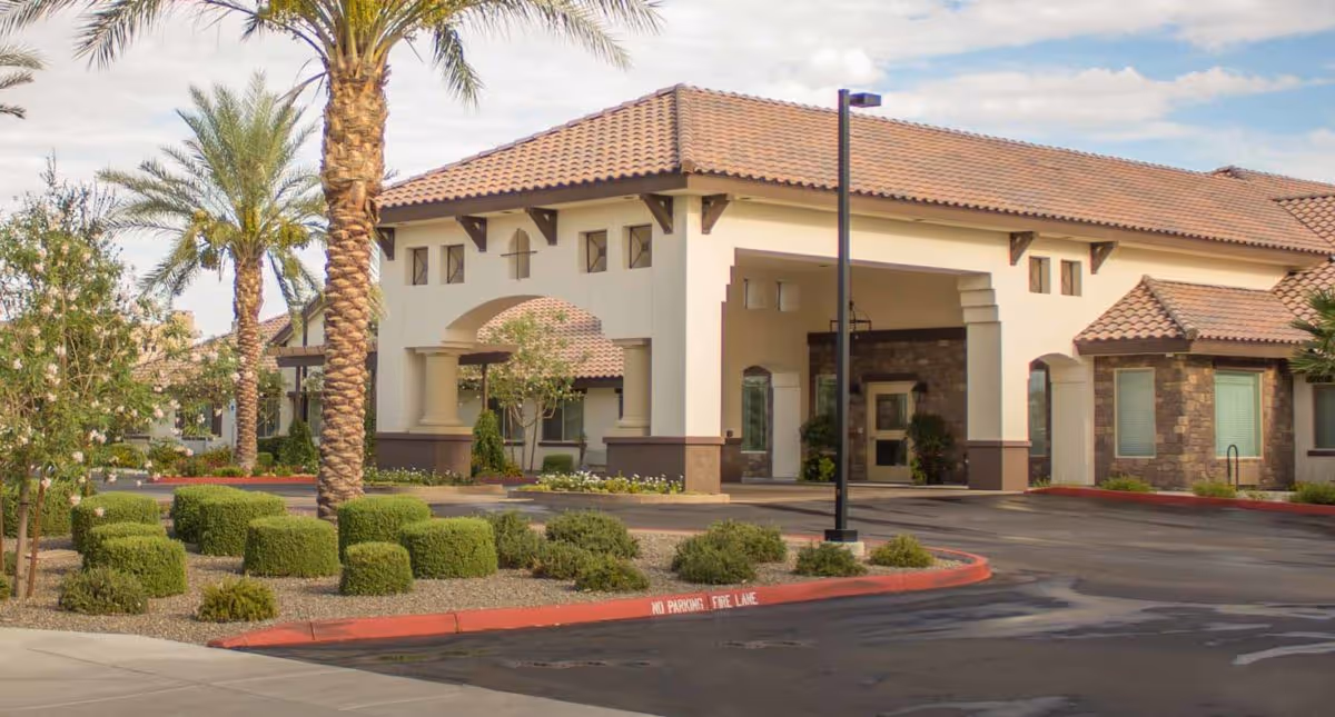 Exterior view of Amber Creek Inn Memory Care Community building with a covered entrance, palm trees, trimmed bushes, and a paved driveway with a no parking fire lane curb.