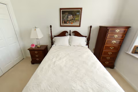 A neatly made bed with white bedding and two pillows in a bedroom. On the left side of the bed is a wooden nightstand with a lamp and a small pink flower decoration. On the right side is a tall wooden dresser with multiple drawers. A framed painting hangs on the wall above the bed. The room has white walls and a window on the right side.