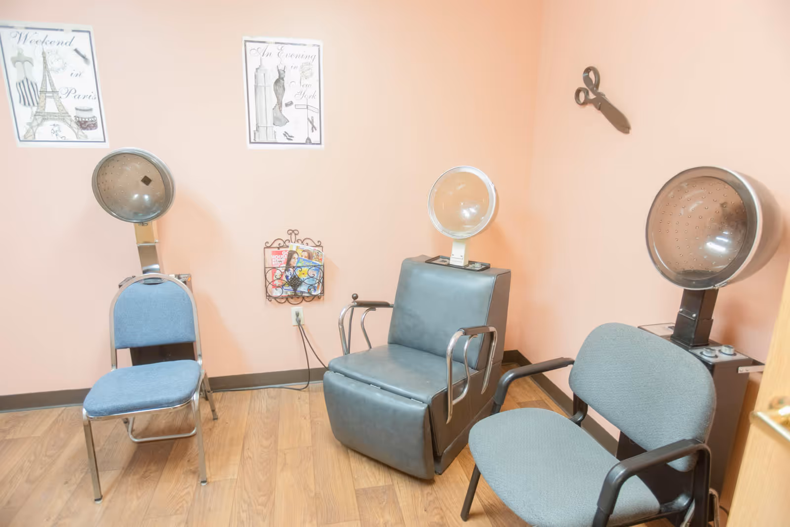 Small salon-style room with three hair-drying chairs and hooded dryers against peach-colored walls and wood flooring.