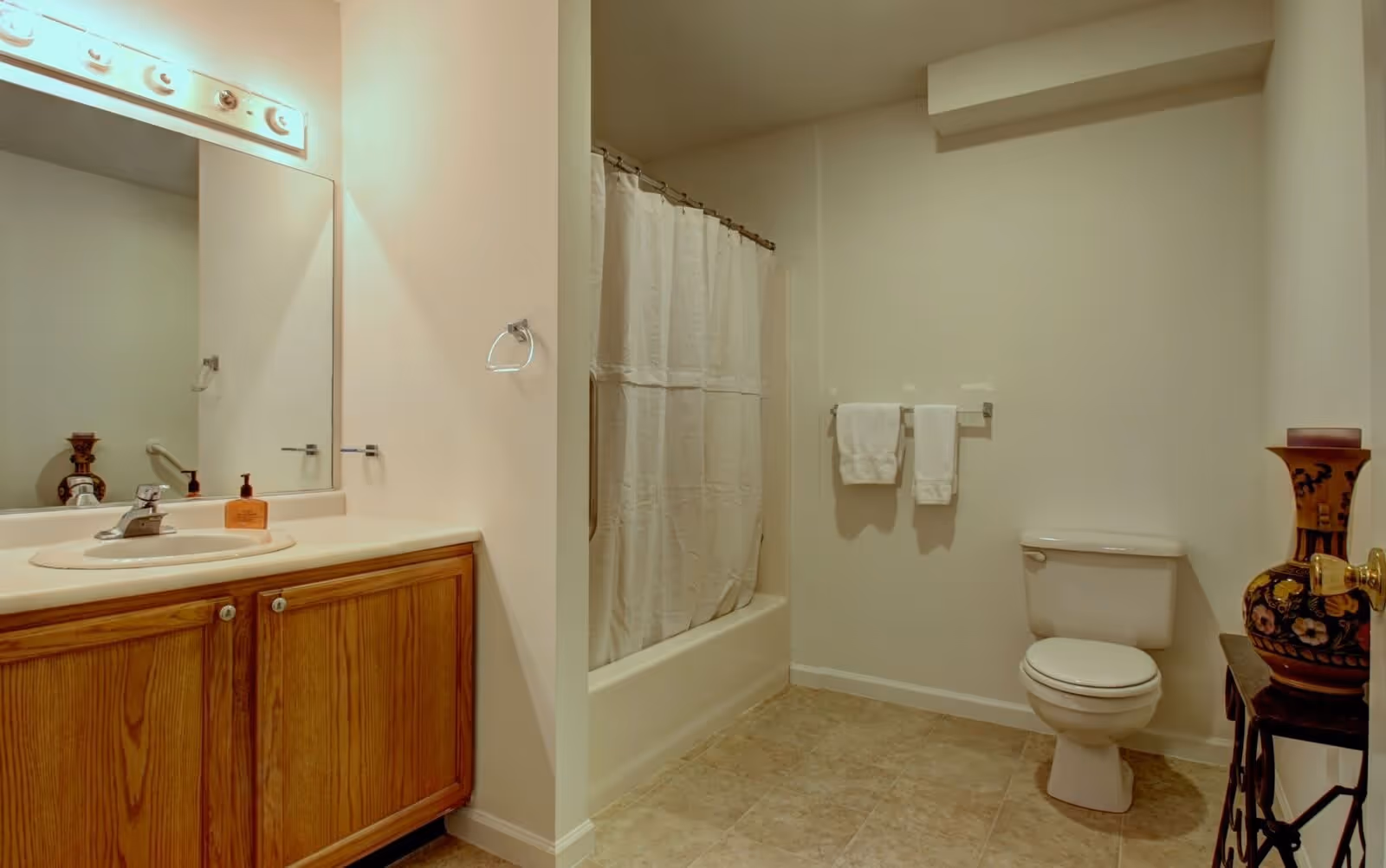 A bathroom with a wooden vanity cabinet, a sink with a faucet, a large mirror with lights above it, a bathtub with a shower curtain, a toilet, two white towels hanging on a towel rack, and a decorative vase on a small table.