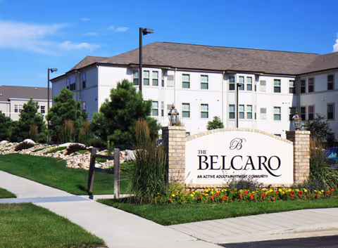 A landscaped entrance sign reading "The Belcaro" in front of a multi-story senior living building under a blue sky.