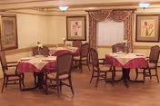 A dining room with several round tables covered with white and burgundy tablecloths, each set with plates, glasses, and napkins. The room has wooden flooring, framed floral artwork on the walls, and a window with draped curtains.