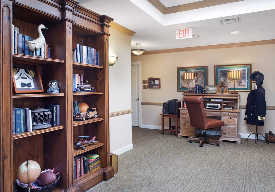 A cozy interior corner of a senior living facility featuring a wooden bookshelf filled with books and decorative items, a wooden desk with a brown leather chair, vintage-style lamps, framed pictures on the wall, and a coat rack holding a pilot's jacket and hat. The carpeted floor and neutral-colored walls create a warm atmosphere.