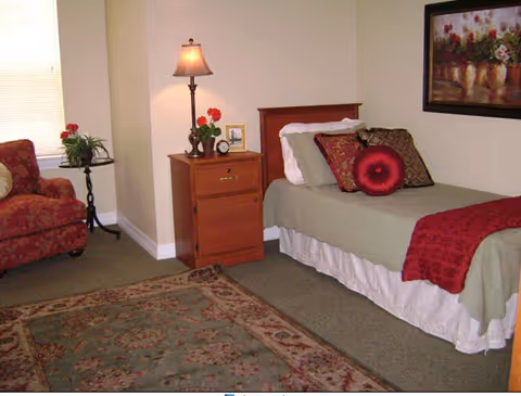 A cozy bedroom in a senior living facility featuring a single bed with green bedding, decorative pillows, and a red throw blanket. Next to the bed is a wooden nightstand with a lamp, a small clock, a framed photo, and a potted plant. There is a patterned armchair near a window with blinds, a small round side table with a plant, and a large decorative rug on the carpeted floor. A framed floral painting hangs on the wall above the bed.