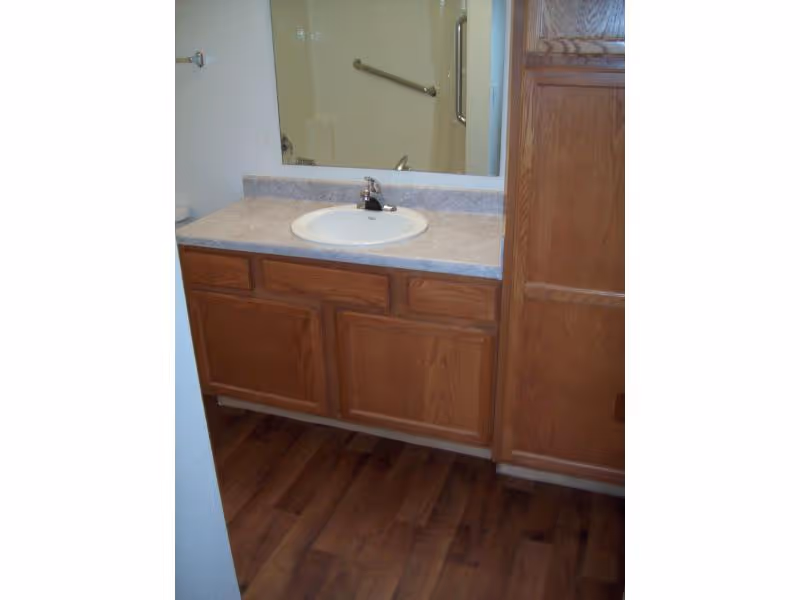 Bathroom vanity with a white sink, silver faucet, and a large mirror above. The countertop is light-colored, and there are wooden cabinets below and to the side. The floor has a wood-like finish.