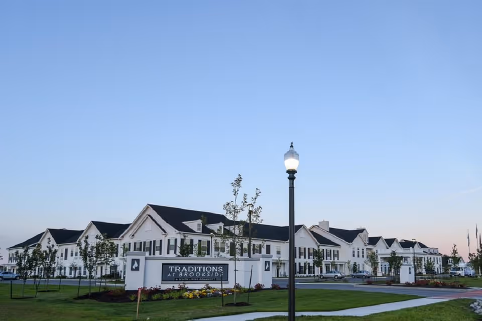 Front exterior of the Traditions at Brookside senior living building with a lawn, entrance sign, and a lit lamppost.