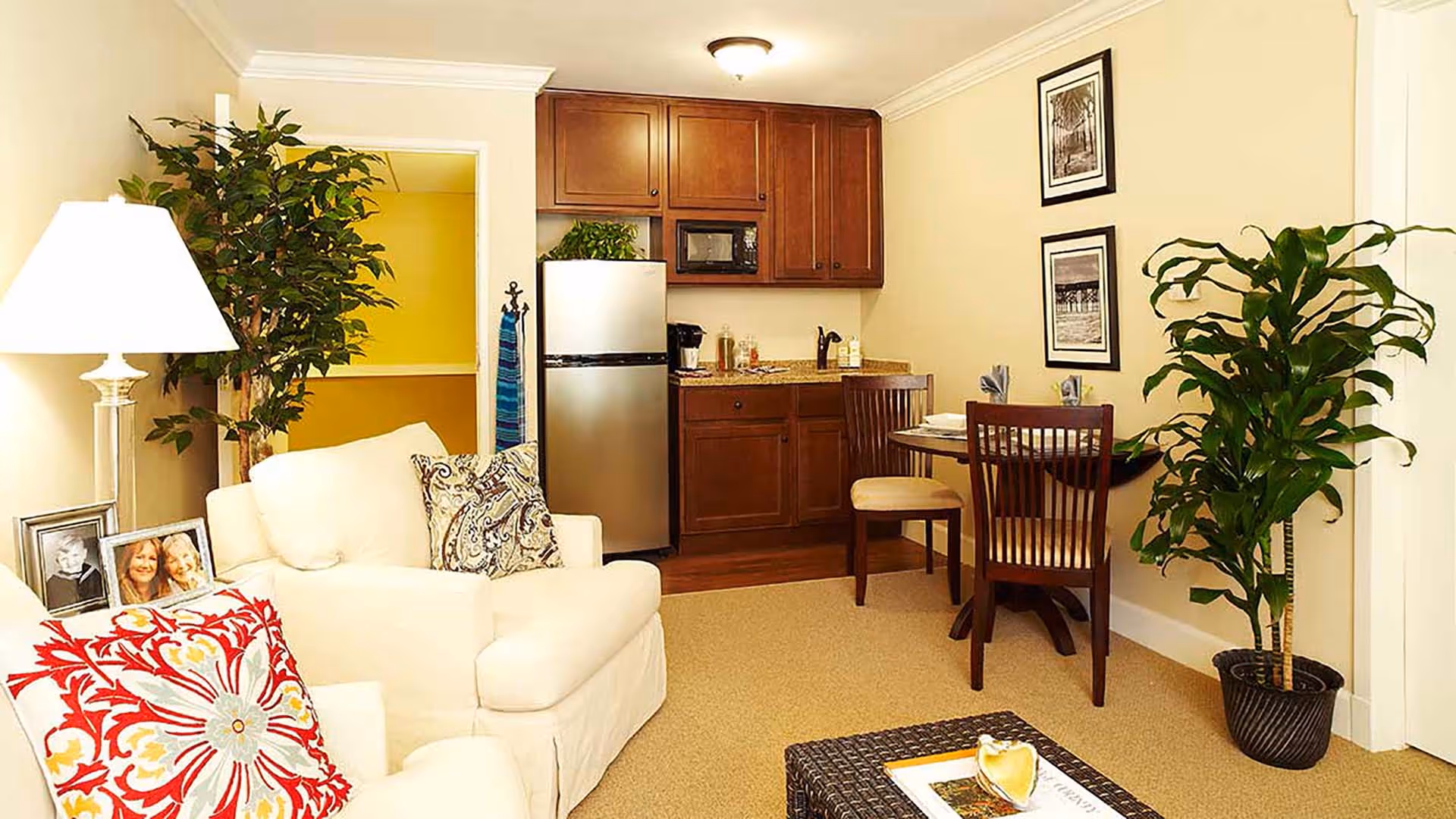 A cozy living area with a white armchair adorned with patterned pillows, a side table with framed photos and a lamp, a small dining table with two chairs, and a kitchenette with wooden cabinets, a stainless steel refrigerator, and a microwave. There are two framed pictures on the wall and two large potted plants adding greenery to the space.