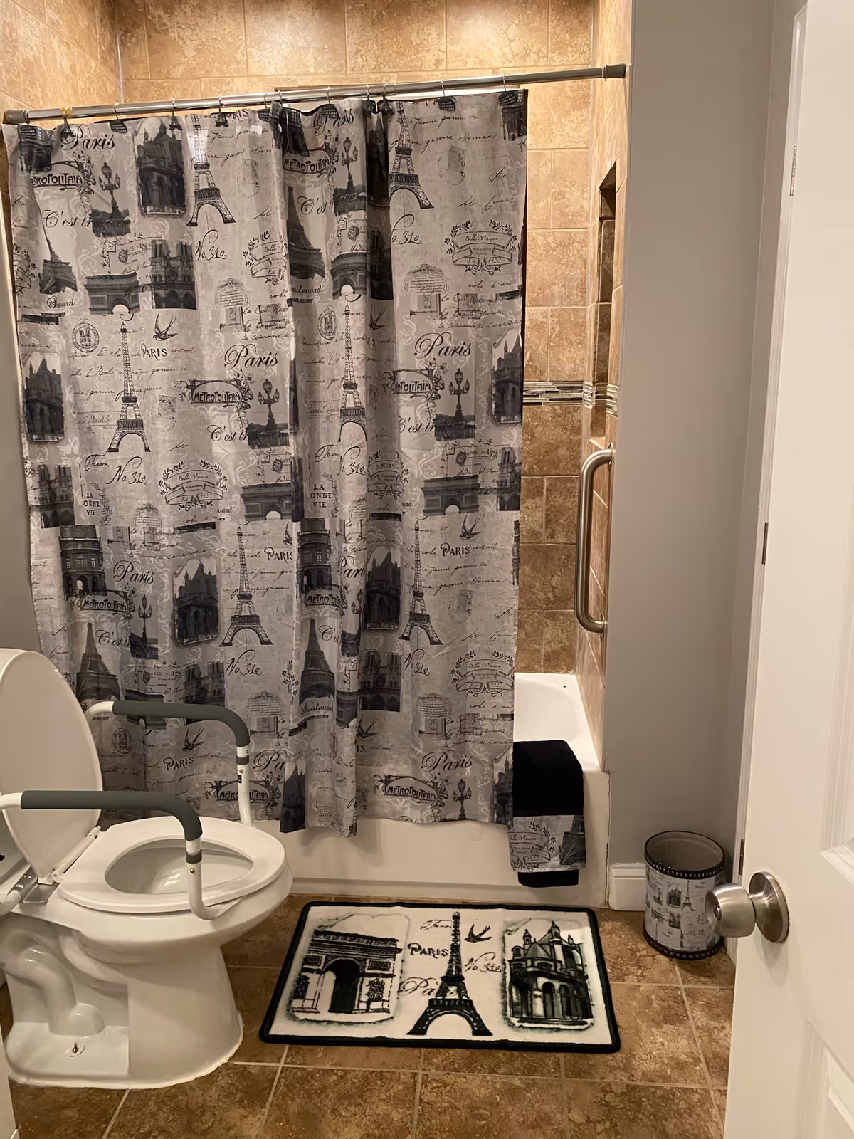 A bathroom with a toilet equipped with safety rails, a bathtub with a Paris-themed shower curtain, a matching floor mat, and a small trash can. The walls and floor are tiled in a beige color.