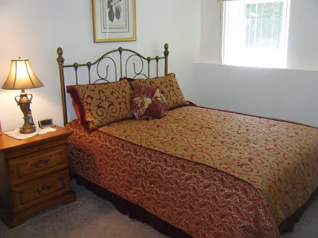 A bedroom with a bed covered in a red and gold patterned bedspread and matching pillows. There is a wooden nightstand with a lamp and a clock on it next to the bed. A framed picture hangs on the wall above the bed, and a window with vertical blinds lets in natural light.