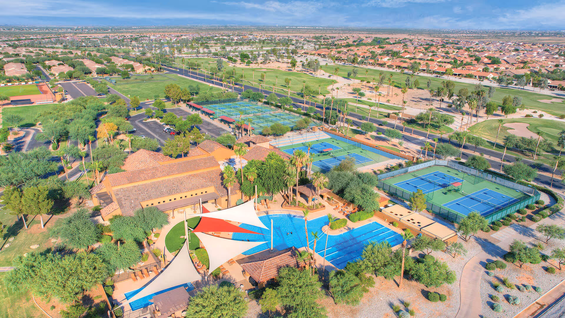 Aerial view of Robson Ranch Arizona showing a large outdoor recreational area with a swimming pool, shaded seating areas, multiple tennis and pickleball courts, surrounded by green lawns, trees, and residential houses in the background under a clear blue sky.