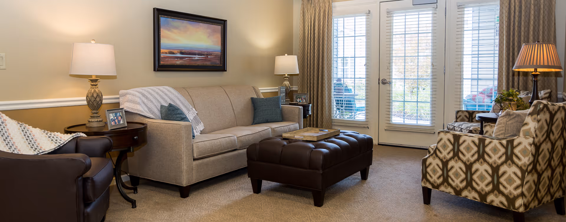 Comfortable living room with a beige sofa, patterned armchairs, a tufted leather ottoman, side tables with lamps, and glass doors to the outside.