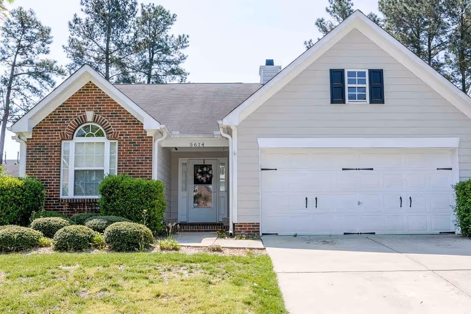 Front exterior of a single-story house with a two-car garage, brick and siding facade, front door, and landscaped shrubs.