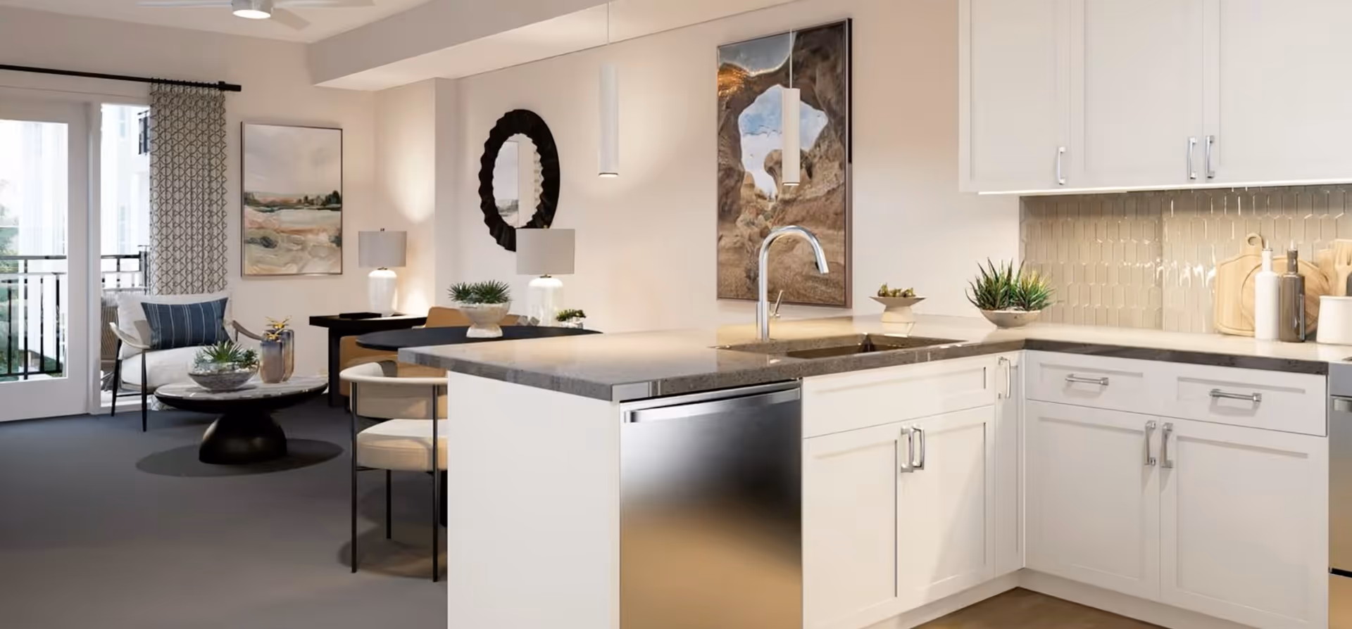 Modern kitchen area with white cabinets, a gray countertop, a stainless steel dishwasher, and a sink with a curved faucet. The kitchen opens to a living space with a round coffee table, a chair with a blue pillow, a small dining table with chairs, decorative plants, a round mirror, and framed artwork on the walls. Large windows with patterned curtains allow natural light into the room.