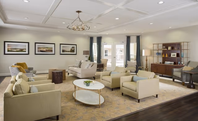 Bright, spacious assisted-living common room with sofas, armchairs, coffee tables, artwork, and shelving.