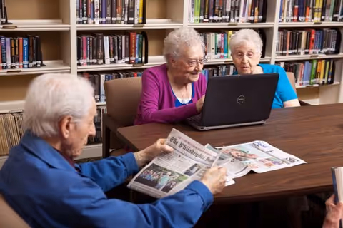 Three elderly individuals sitting at a wooden table in a library or reading room. Two women are looking at a laptop screen, while a man is reading a newspaper. Bookshelves filled with books are visible in the background.