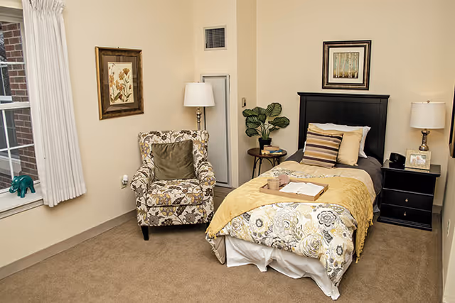 A cozy bedroom in a senior living facility featuring a single bed with floral and yellow bedding, a black headboard, and multiple pillows. Next to the bed is a black nightstand with a lamp, a framed photo, and a clock. A small round table with a plant and books is beside the bed. There is a patterned armchair with a green cushion near a window with white curtains. The walls are decorated with framed floral artwork.