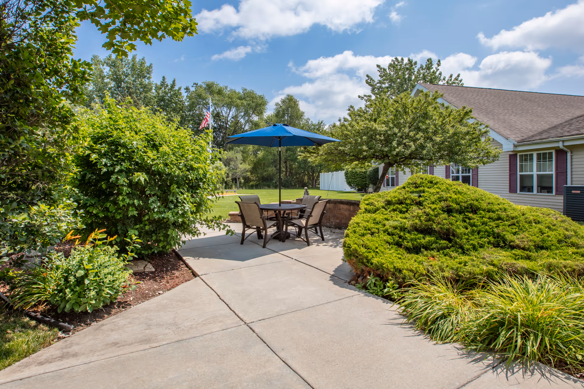Outdoor patio area with a round table and four chairs under a blue umbrella, surrounded by green bushes and trees, with a building and a partly cloudy sky in the background.