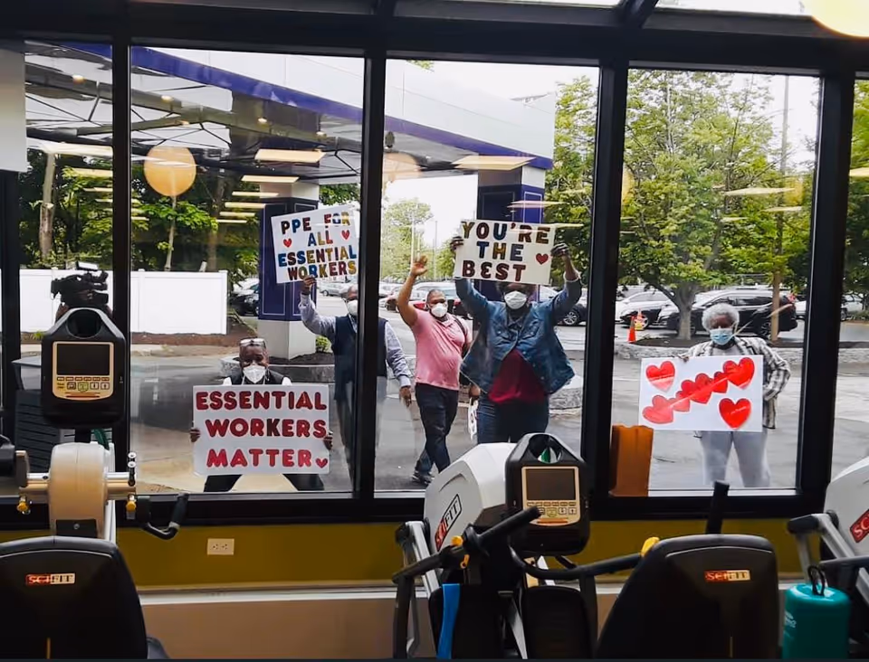 View through large windows of several people outside holding signs that say 'PPE for all essential workers', 'You're the best', 'Essential workers matter', and a sign with red hearts. Inside, exercise equipment is visible in the foreground.