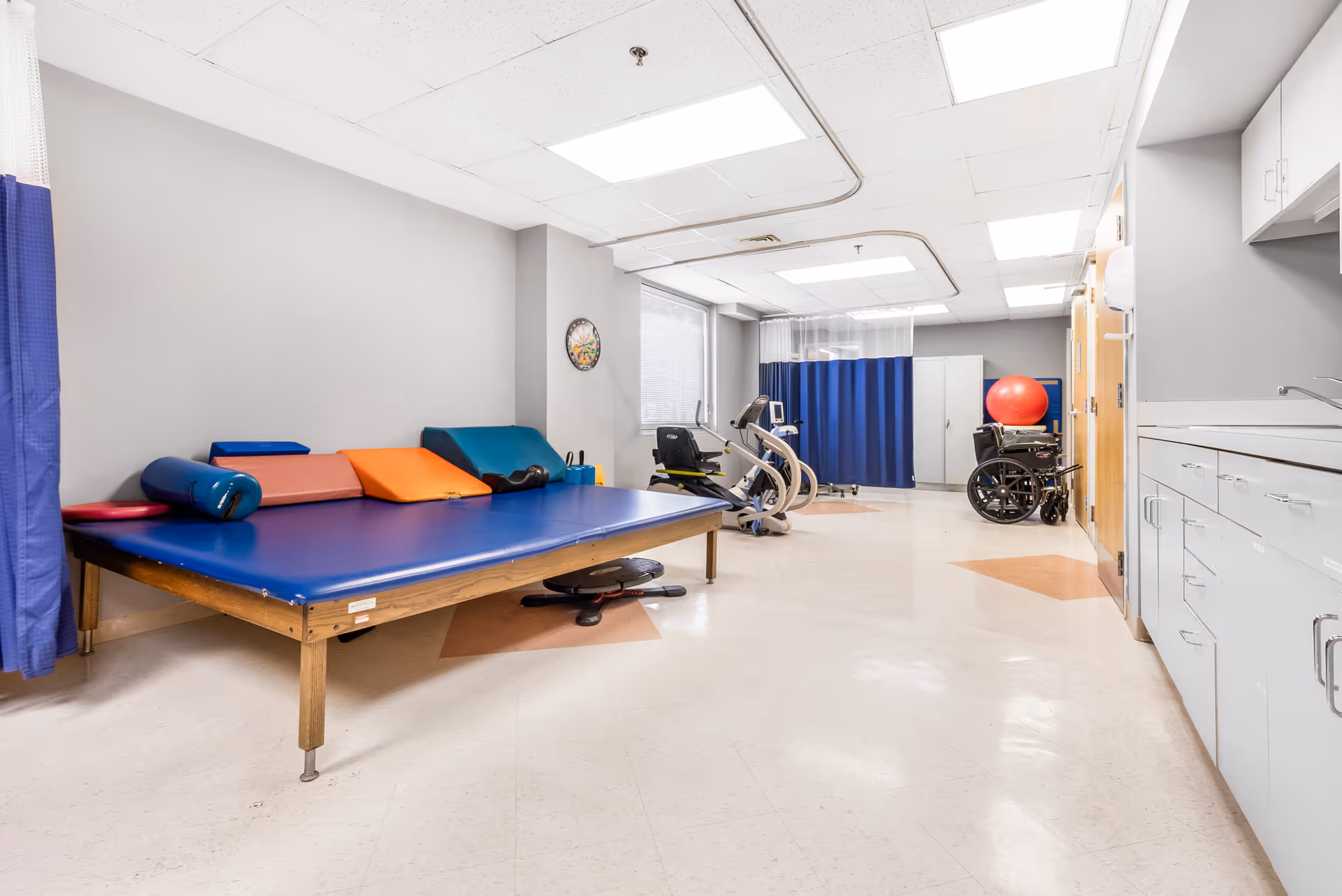 A rehabilitation therapy room with a large padded therapy table covered in blue vinyl and various colored cushions. The room has exercise equipment including a stationary bike and a wheelchair. There is a red exercise ball near the back, a blue privacy curtain, and cabinets with a sink along the right wall. The floor is tiled, and the ceiling has fluorescent lighting.
