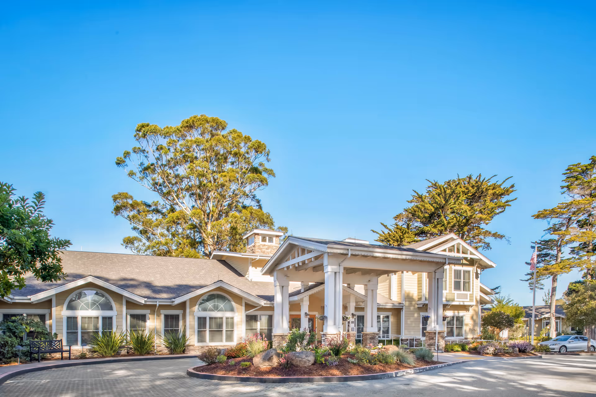Front exterior of a senior living facility with a covered porte-cochere entrance, landscaped driveway, and large trees.