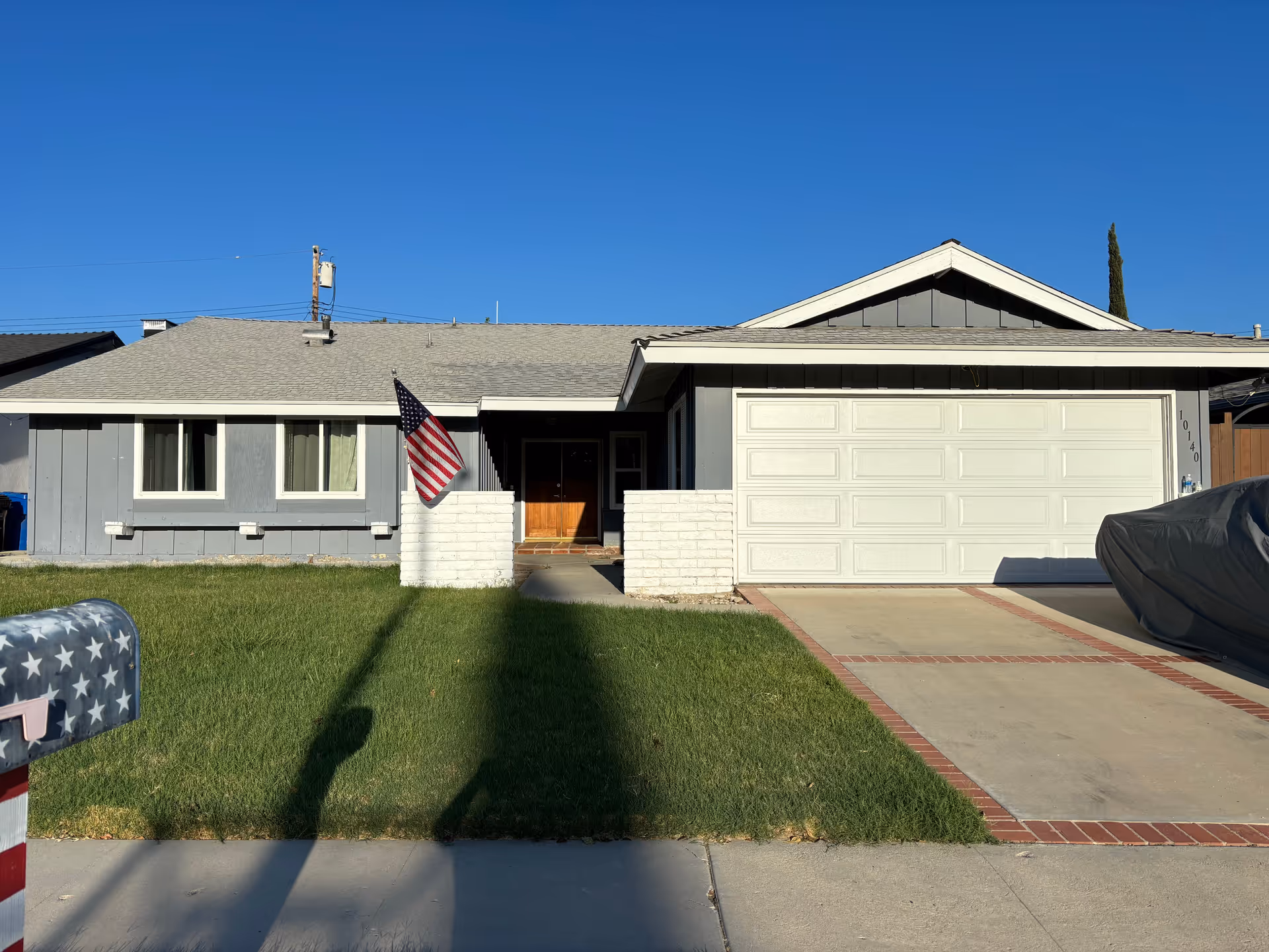 Single-story residential house with a gray exterior, white garage door, and a small front lawn. An American flag is mounted on a white brick pillar near the entrance. The sky is clear and blue.