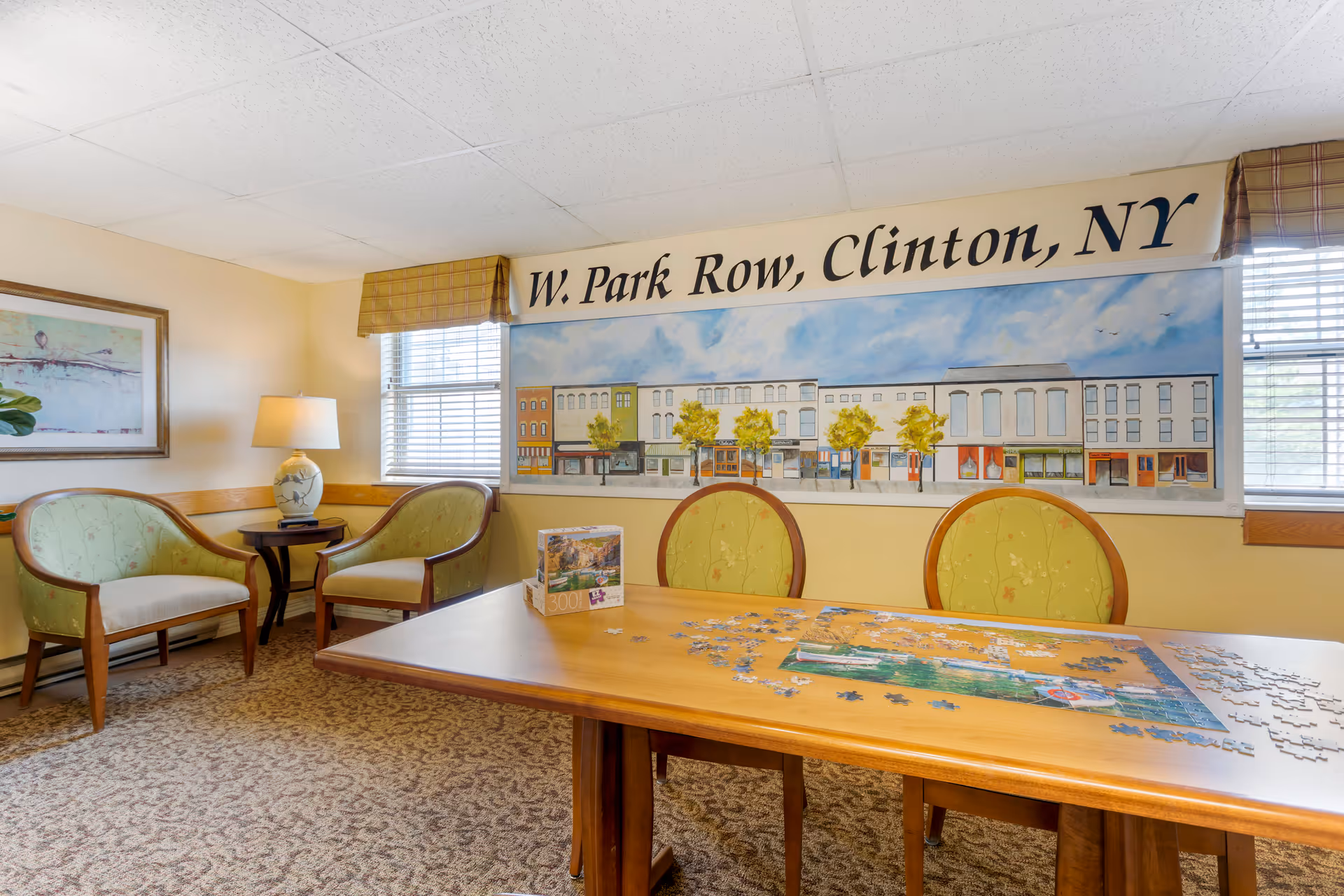A cozy room with a wooden table in the center featuring a partially completed jigsaw puzzle. Two green cushioned chairs are placed at the table. In the background, there are two green armchairs with wooden frames, a side table with a lamp, and a framed artwork on the wall. A large mural depicting a street scene with buildings and trees is displayed on the wall, with the text 'W. Park Row, Clinton, NY' above it. The room has carpeted flooring and windows with blinds.