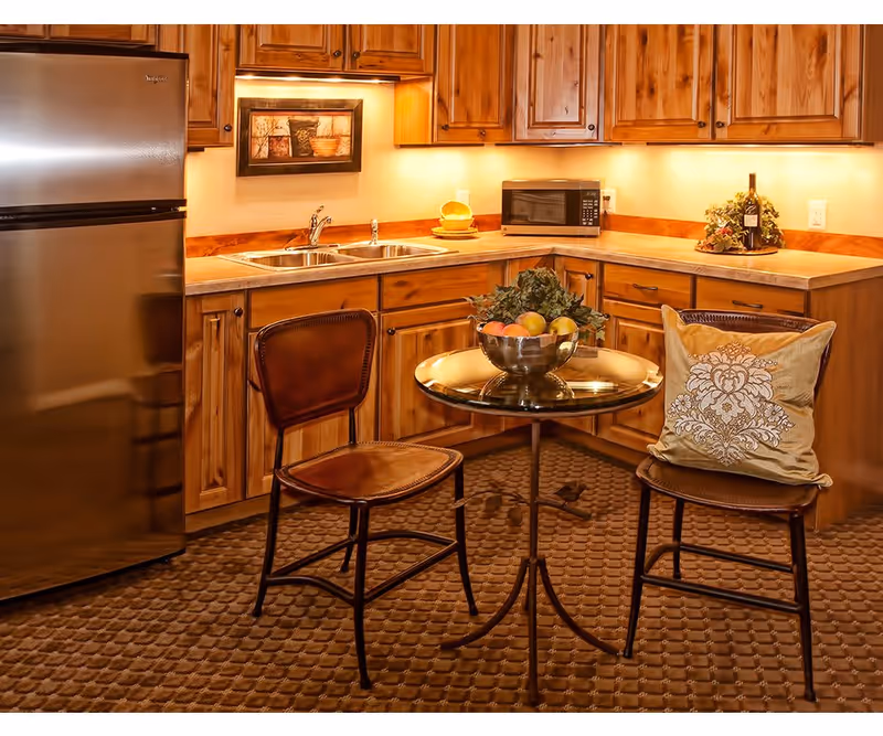 A cozy kitchen area with wooden cabinets, a stainless steel refrigerator, a double sink, a microwave, and a small round table with two chairs. One chair has a decorative pillow, and a bowl of fruit is on the table. The countertop has a bottle of wine and a small plant. The lighting under the cabinets creates a warm ambiance.
