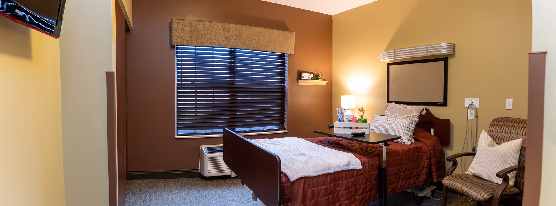 A cozy bedroom in a senior living facility featuring a single bed with a brown quilt and white blanket, a bedside table with a lamp and decorative items, a cushioned armchair with a white pillow, a window with closed blinds, and a wall-mounted television.