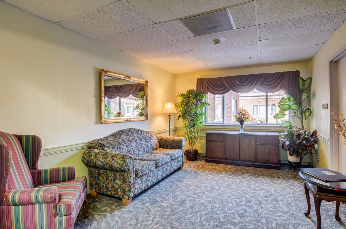 A cozy common room with a patterned sofa and striped armchair, window with a valance, plants, lamp, and a wooden cabinet.