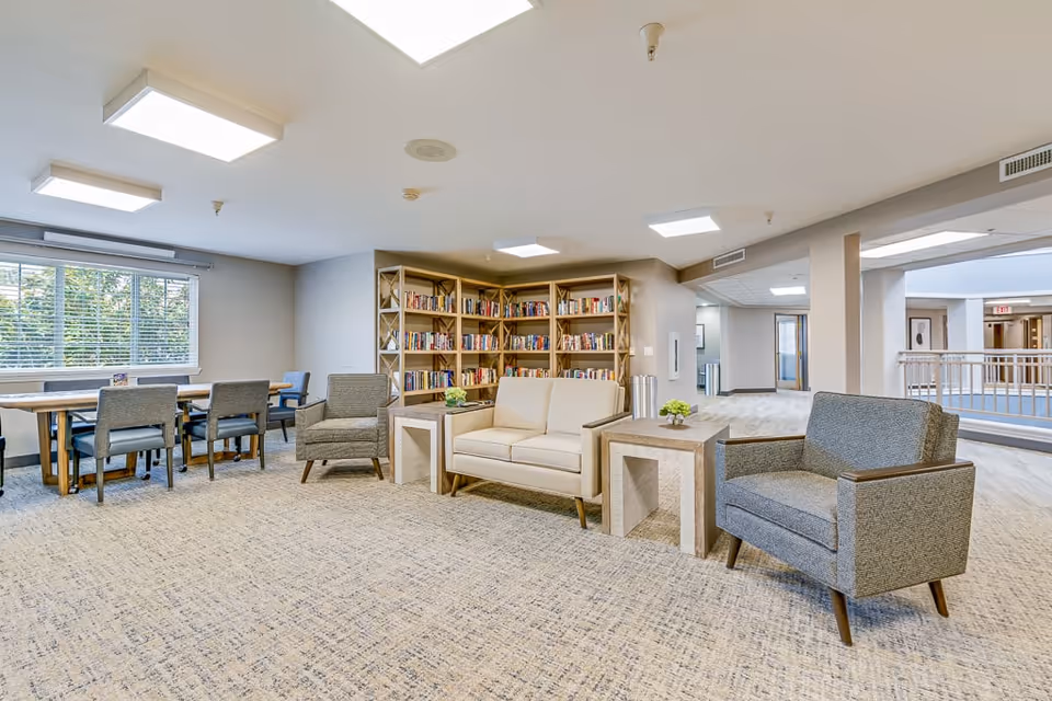 A bright and spacious common area in an assisted living facility featuring a seating arrangement with two armchairs and a loveseat around two small tables with plants. In the background, there is a wooden bookshelf filled with books and a table with several chairs near a window with blinds. The area has carpeted flooring and overhead lighting.