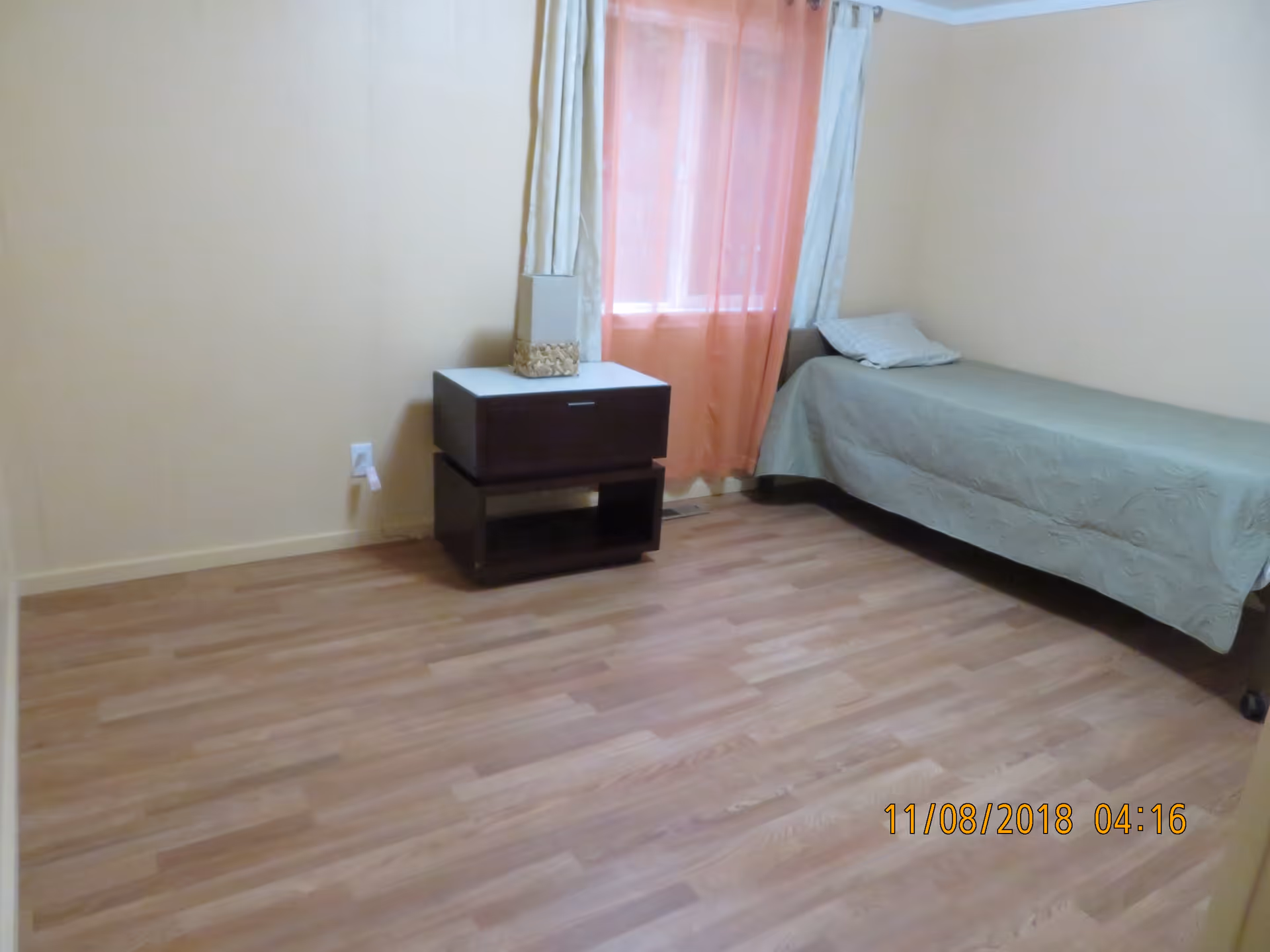 Sparsely furnished bedroom with a single bed, nightstand with a lamp, and wood-look flooring.
