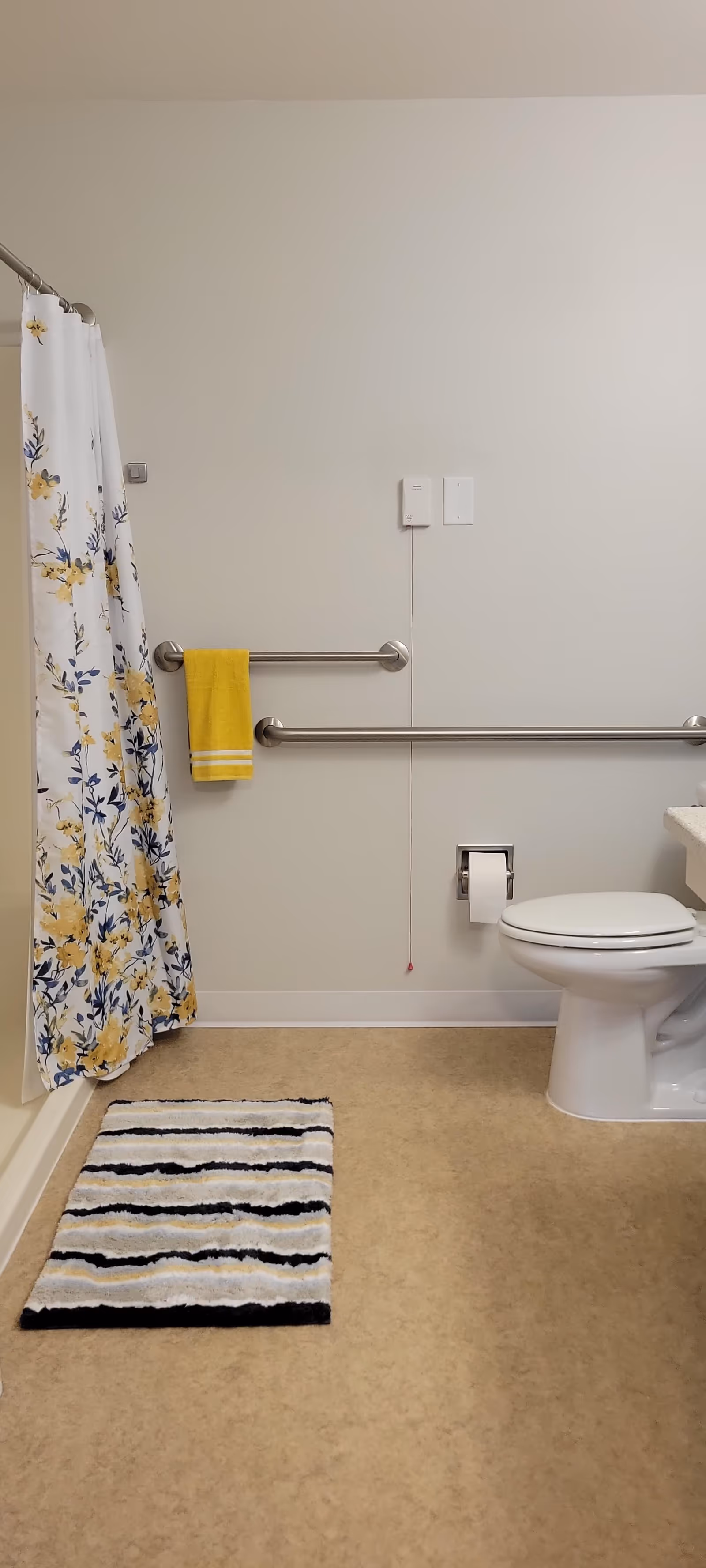 A bathroom with a white toilet, a floral shower curtain with yellow and blue flowers, a yellow towel hanging on a metal grab bar, and a striped bath mat on the floor. The walls are light-colored and there are two metal grab bars installed horizontally on the wall.