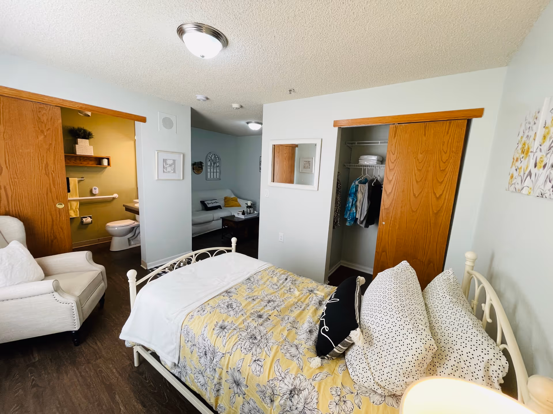 A cozy bedroom in a senior living facility with a bed covered in a yellow floral comforter and multiple pillows. To the left, there is a white armchair and an open door leading to a bathroom with a toilet and grab bars. On the right side, there is an open closet with clothes hanging inside. In the background, a small living area with a white couch and a coffee table is visible. The walls are painted light blue, and the floor is dark wood.