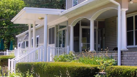 Exterior view of a nursing and rehabilitation center building with a covered porch supported by white columns, surrounded by green bushes and plants.