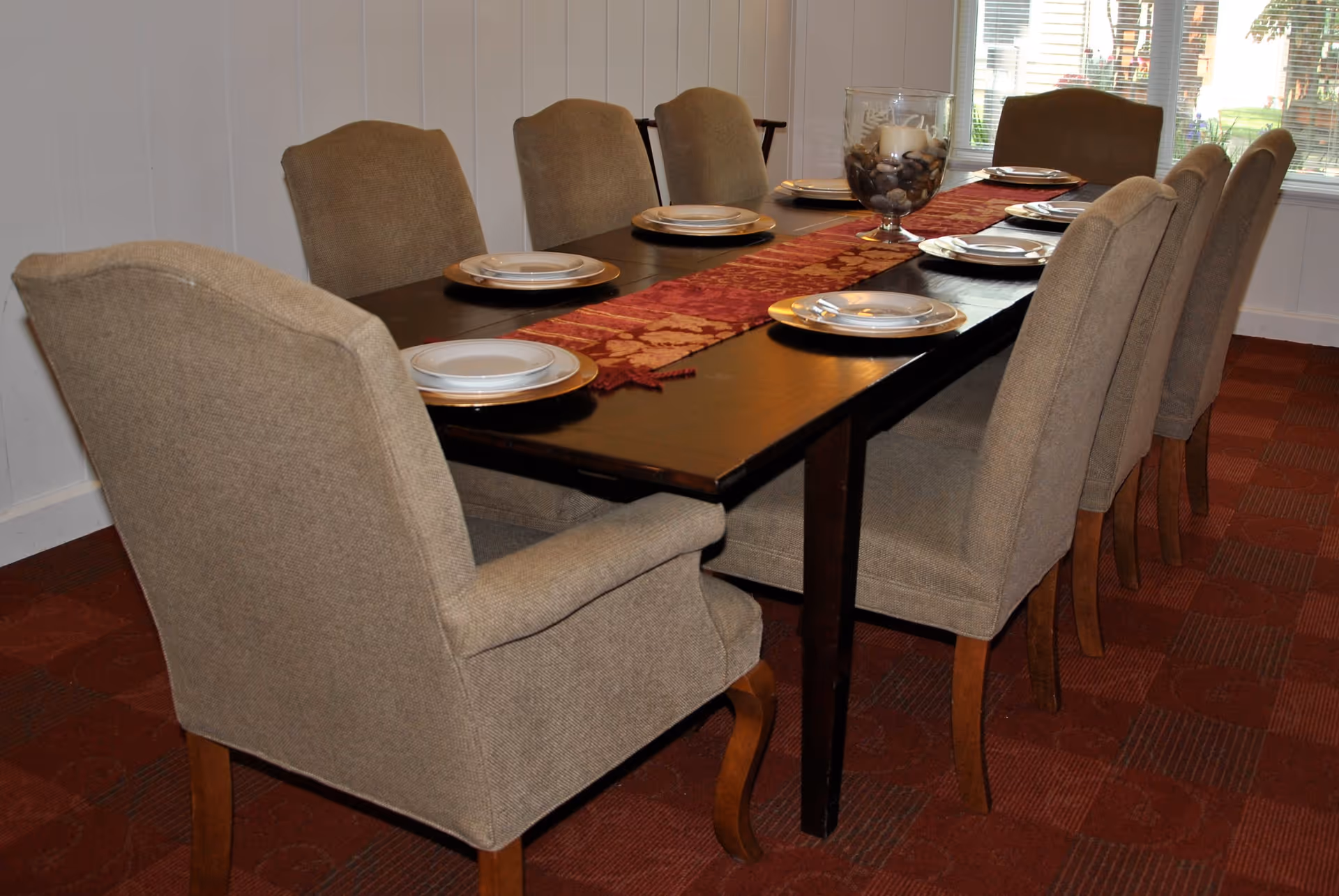 A dining room with a long dark wooden table set with eight beige upholstered chairs. The table is decorated with a red and gold patterned runner and a centerpiece consisting of a glass container filled with rocks and a candle. Plates and silverware are neatly arranged at each place setting. A window with blinds is visible in the background.