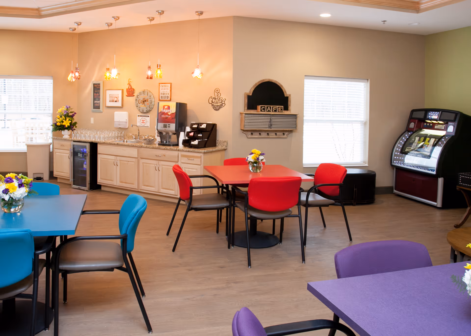 Community dining/activity room with colorful tables and chairs, a kitchenette counter, and a jukebox.
