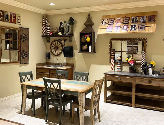A cozy interior room with a rustic wooden dining table surrounded by five chairs. On the left wall, there is a decorative window frame with shutters and a sign that reads 'CAFE'. The back wall features a small sink with a countertop, shelves holding decorative items, and a paper towel dispenser. Above the sink, a wooden sign reads 'GENERAL STORE'. To the right, there is a wooden sideboard with decorative items including flowers and striped candle holders, and a mirror hanging above it. The room has beige walls and light-colored flooring.