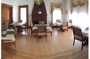 A dining room with several wooden tables and chairs arranged neatly. The room features large windows with decorative curtains allowing natural light to fill the space. There is a dark wooden fireplace with a mantel on one wall, and the floor is made of light-colored wood.