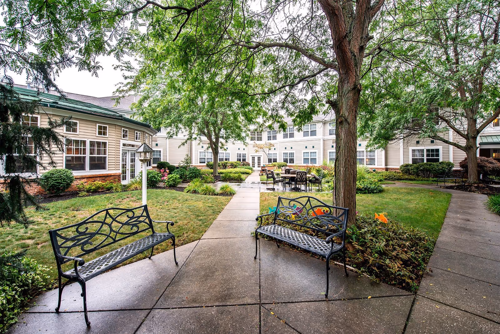 Outdoor courtyard area at Westlake Woods Assisted Living featuring two black metal benches, a paved walkway, green grass, trees, and shrubs with the building surrounding the courtyard.