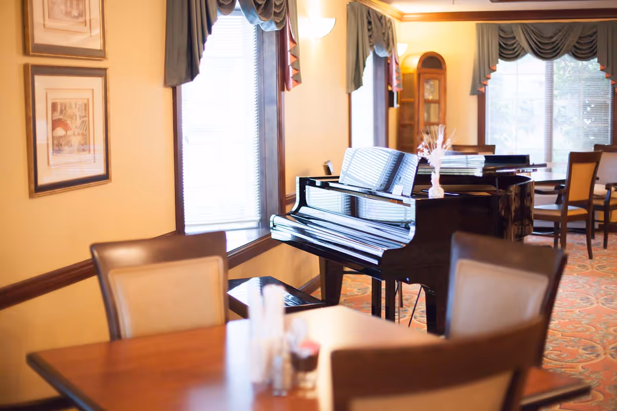 A bright dining room with a glossy black grand piano, tables and chairs, and windows with draped valances.