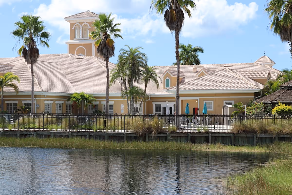 Beige Mediterranean-style senior living building with palm trees, a fenced patio, and a reflecting pond in the foreground.