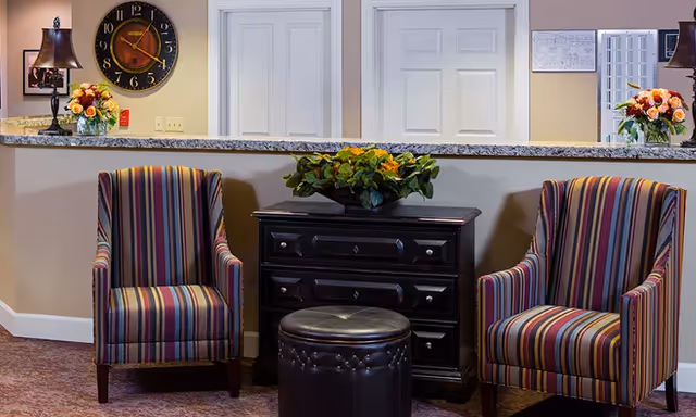 A reception area with a granite countertop behind which are two closed white doors. In front of the counter are two striped armchairs with a small black chest of drawers and a round black ottoman between them. There are flower arrangements on the counter and on the chest of drawers, a table lamp, a large wall clock, and a bulletin board on the wall.