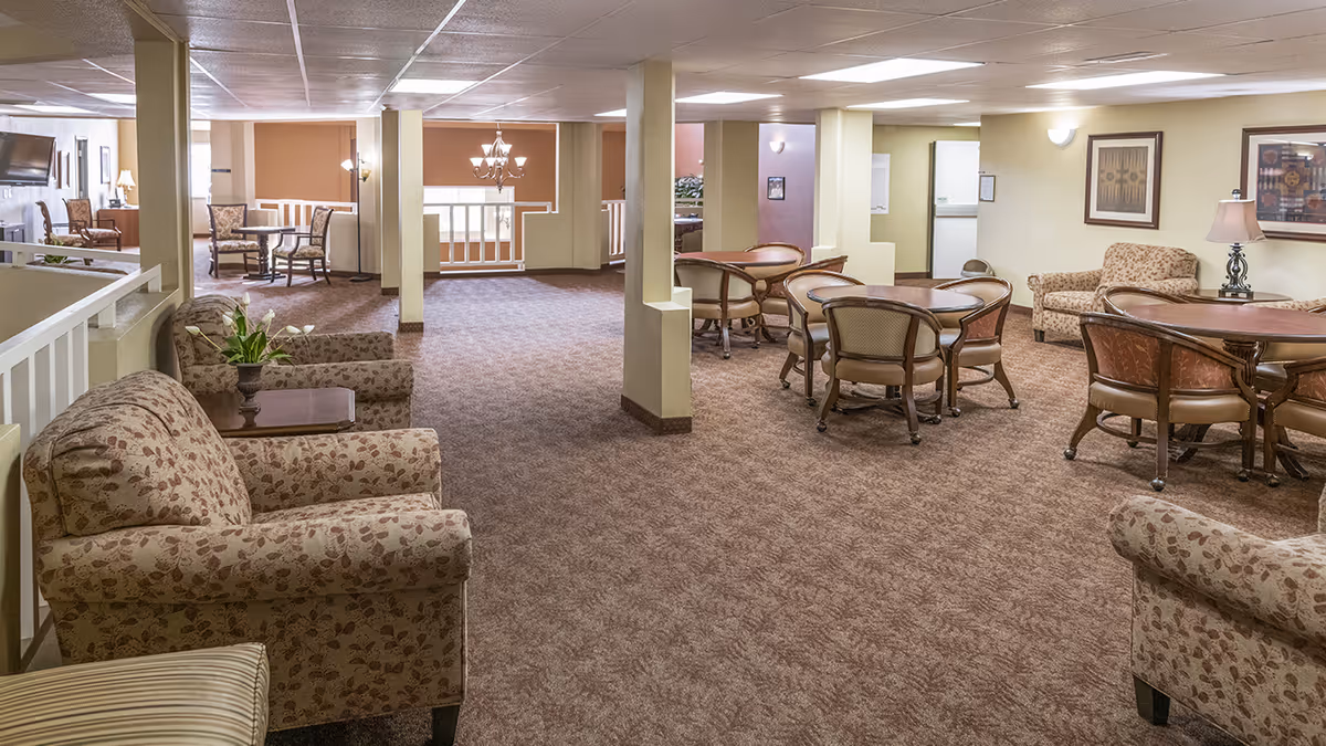 A spacious senior living facility common area with patterned armchairs, round tables with chairs, framed artwork on the walls, and soft lighting. The room has carpeted floors and beige walls, with a chandelier visible in the background near a railing.