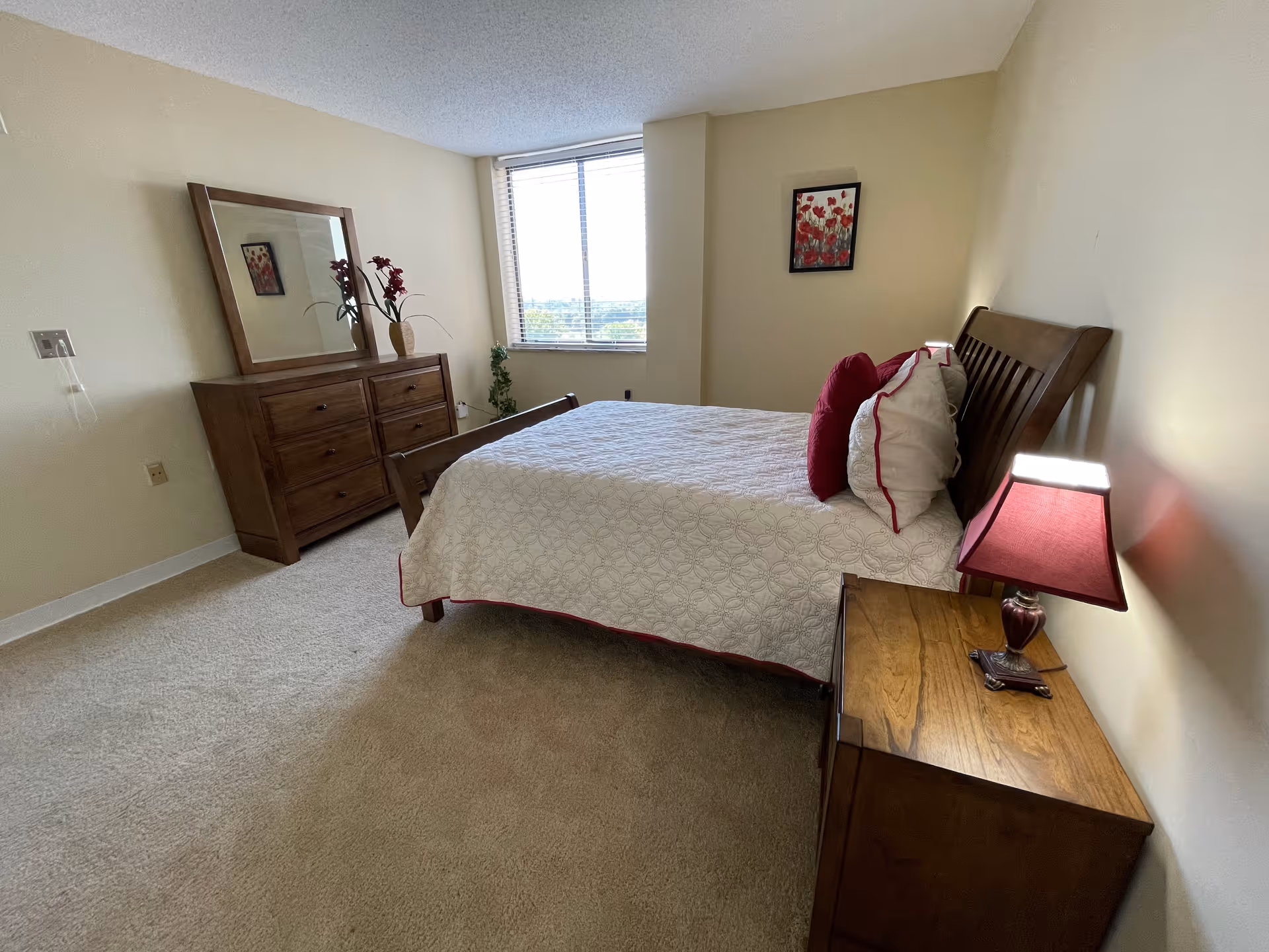 A bedroom with a wooden bed frame and a white bedspread with red trim. There are red and white pillows on the bed. To the right of the bed is a wooden nightstand with a red lampshade lamp. On the left side of the room is a wooden dresser with a large mirror and a vase with red flowers. A window with blinds is centered on the far wall, letting in natural light. A framed picture with red flowers hangs on the wall above the bed.