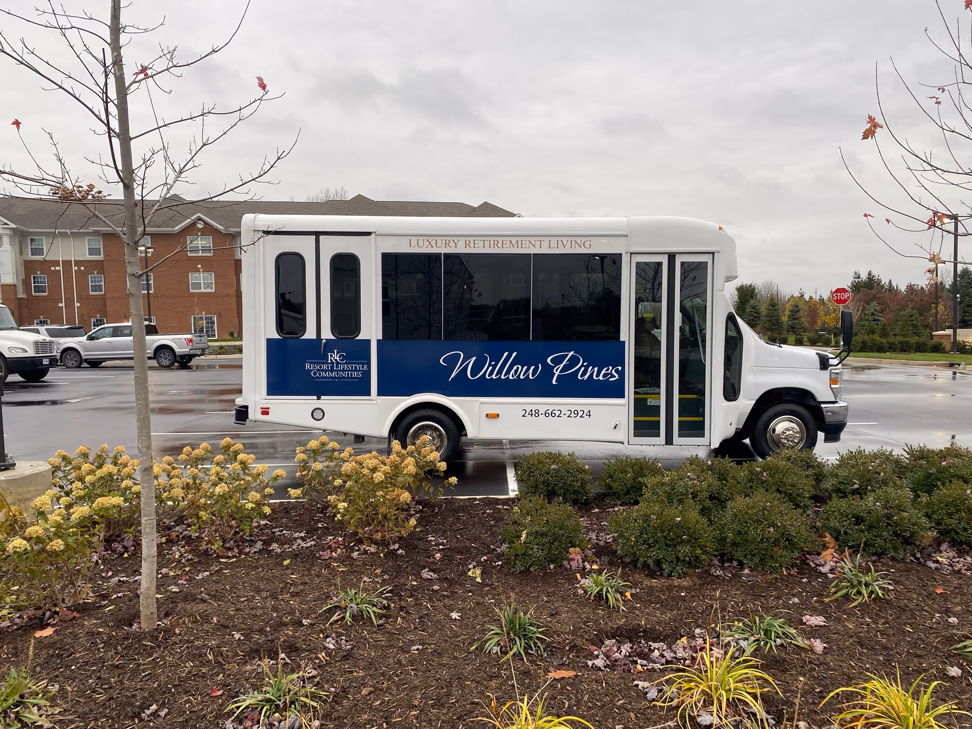 A white shuttle bus labeled "Willow Pines" parked in a lot outside a brick retirement building on a cloudy day.