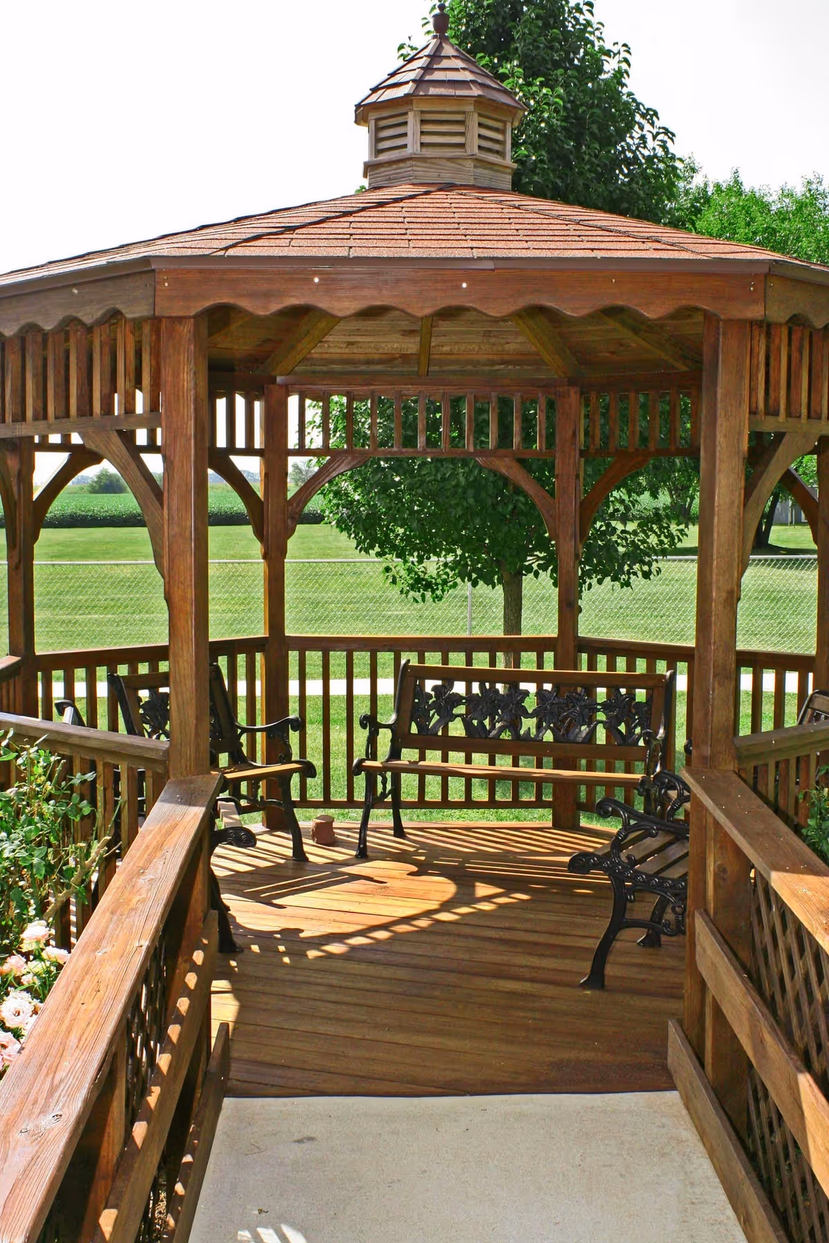 A wooden gazebo with a shingled roof and decorative railing, containing metal benches with ornate backs, situated outdoors with green grass and trees visible in the background.
