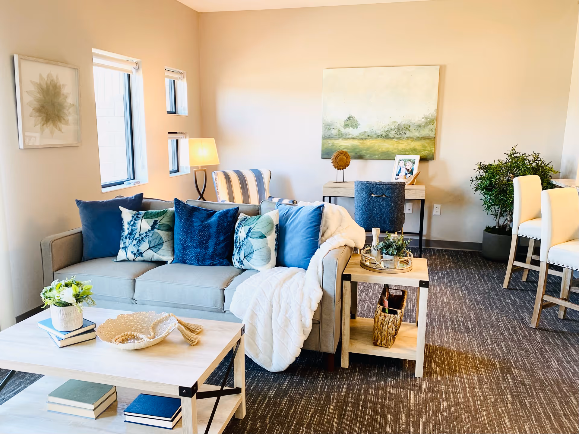 Bright furnished living room with a gray sofa, blue and patterned pillows, coffee and side tables, artwork, and dining chairs.