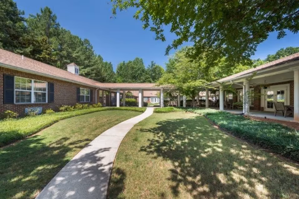 A sunny outdoor courtyard area at Brookstone Alzheimer's Special Care Center with a curved concrete pathway leading through a grassy lawn surrounded by single-story brick buildings with white columns and large windows. Trees provide shade and greenery around the area.